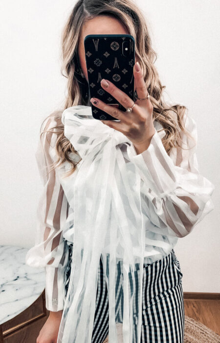 Młoda kobieta prezentuje białą transparentną koszulę z kokardą i spodnie eleganckie w kratę